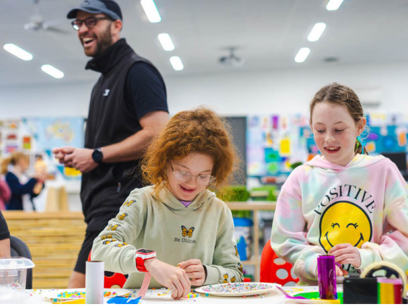 Two girls sitting at table crafting with OSHC educator smiling in the background